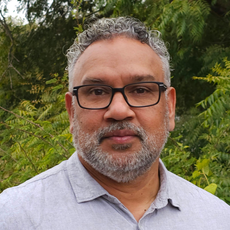 This is an image of Mark Yettica-Paulson. Mark is wearing glasses and a light grey coloured shirt. Mark is standing in front some greenery.